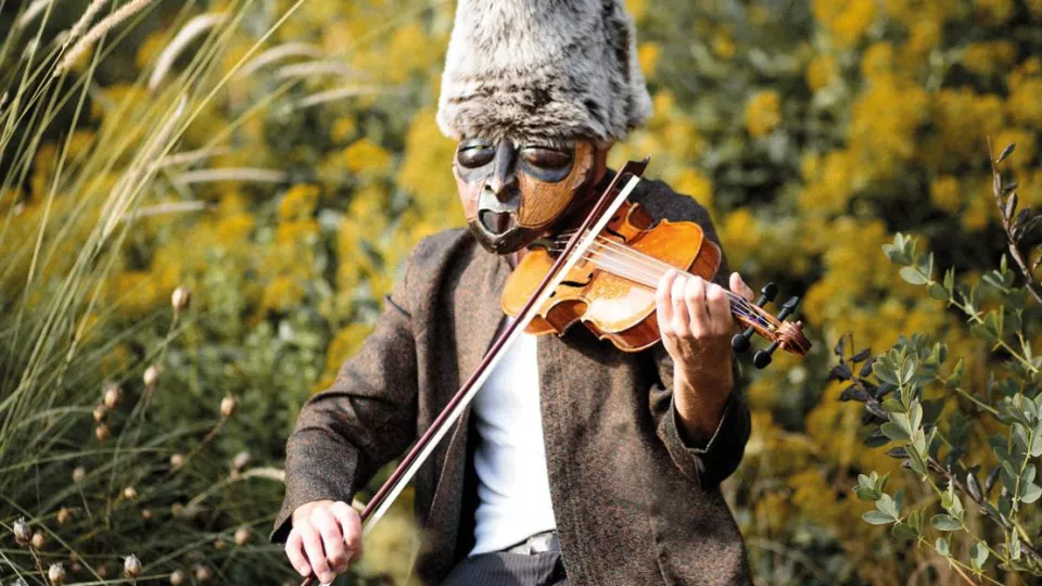 Eine Person mit einer Maske vor dem Gesicht und einem hohen Fellhut auf dem Kopf sitzt in einer Wiese und spielt auf einer Violine