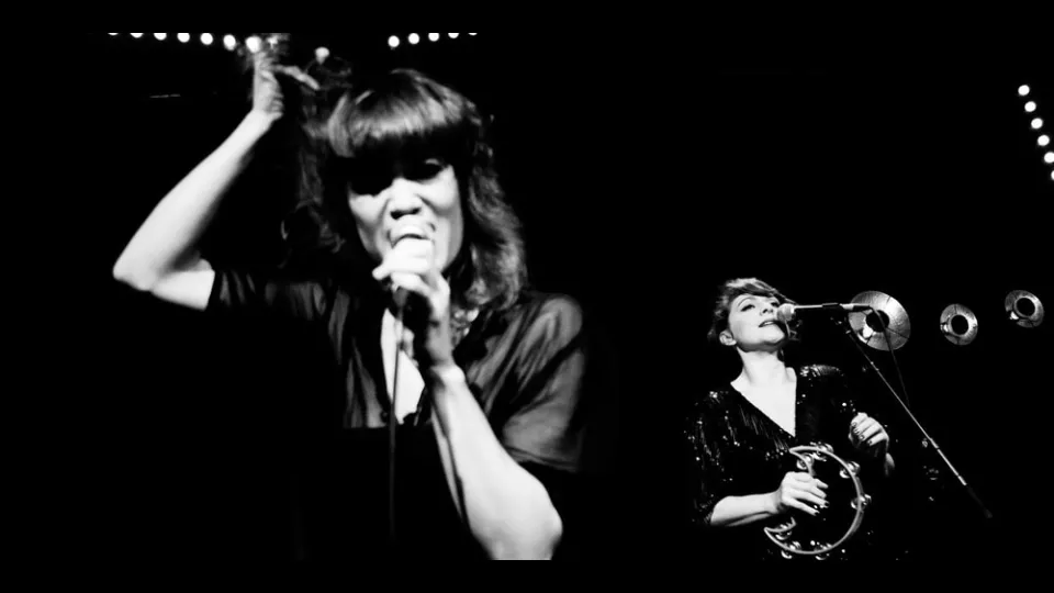 Bandfoto Nouvelle Vagues. Zwei Frauen, jeweils mit Mikrofon. Eine spielt zusätzlich einen Schellenring. Foto in schwarz-grau.